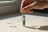 A silver-colored metal pendulum with a green Aventurine stone attached to the end, suspended by a chain.